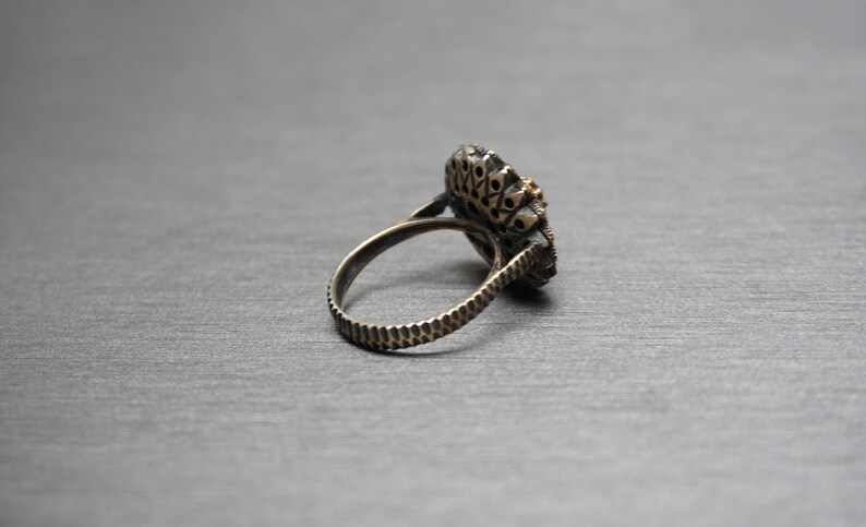 Pu&ograve; includere: Un anello d'argento con un grande designo ornato nero e oro. L'anello &egrave; posto su una superficie grigia.