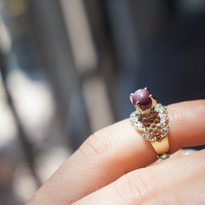 May include: A gold ring with a red gemstone set in a delicate, heart-shaped design. The ring is adorned with small, clear gemstones.