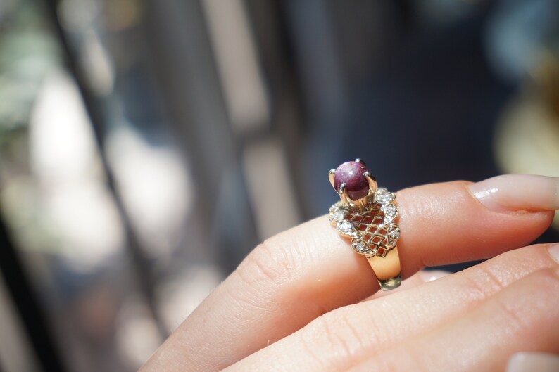 May include: A gold ring with a red gemstone set in a delicate, heart-shaped design. The ring is adorned with small, clear gemstones.
