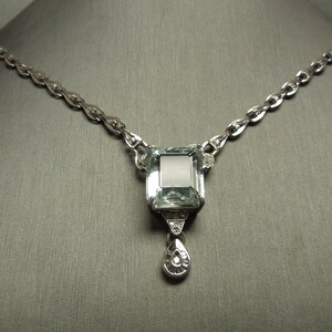 K&ouml;nnte beinhalten: Eine silberne Halskette mit einem gro&szlig;en, rechteckigen, hellblauen Edelsteinanh&auml;nger. Der Anh&auml;nger ist mit kleineren Diamanten und einem tropfenf&ouml;rmigen Anh&auml;nger verziert. Die Kette hat ein filigranes, verbundenes Design, und die Halskette wird vor einem grauen Hintergrund pr&auml;sentiert.