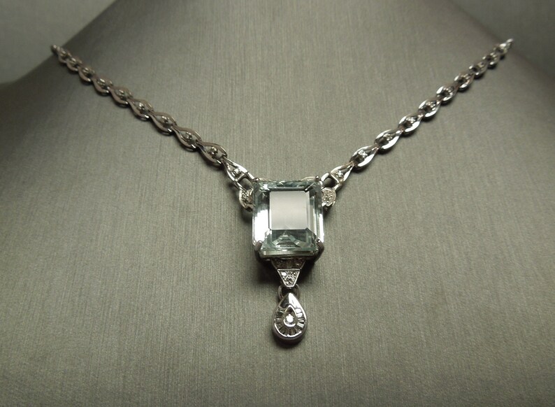 K&ouml;nnte beinhalten: Eine silberne Halskette mit einem gro&szlig;en, rechteckigen, hellblauen Edelsteinanh&auml;nger. Der Anh&auml;nger ist mit kleineren Diamanten und einem tropfenf&ouml;rmigen Anh&auml;nger verziert. Die Kette hat ein filigranes, verbundenes Design, und die Halskette wird vor einem grauen Hintergrund pr&auml;sentiert.