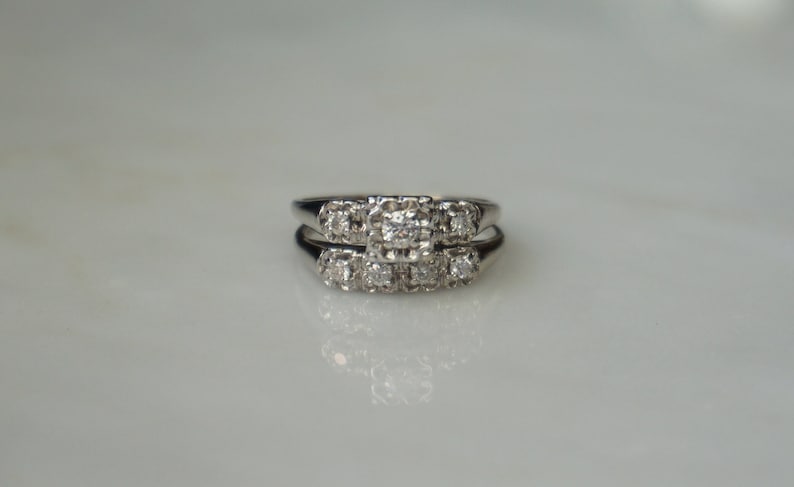 May include: A set of two matching rings, likely a wedding set, with a silver-colored band and multiple small, clear gemstones. The rings are set on a white surface, reflecting light and creating a bright, elegant appearance.