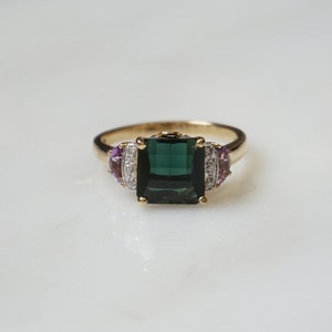 Pu&ograve; includere: Un anello d'oro con una grande pietra preziosa quadrata verde scuro circondata da pietre preziose pi&ugrave; piccole, chiare e viola.