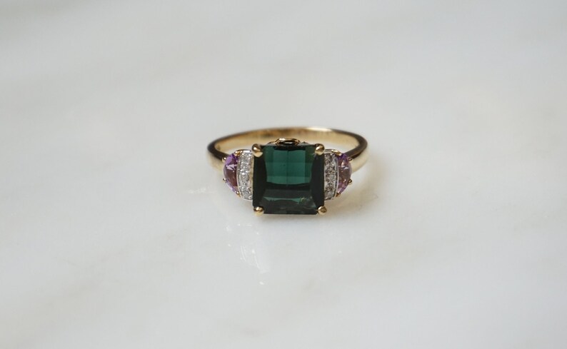 Pu&ograve; includere: Un anello d'oro con una grande pietra preziosa quadrata verde scuro circondata da pietre preziose pi&ugrave; piccole, chiare e viola.