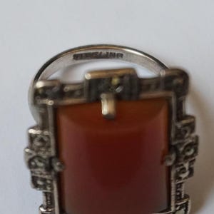 May include: A silver ring with a rectangular carnelian stone set in a marcasite frame. The ring is marked "STERLING".