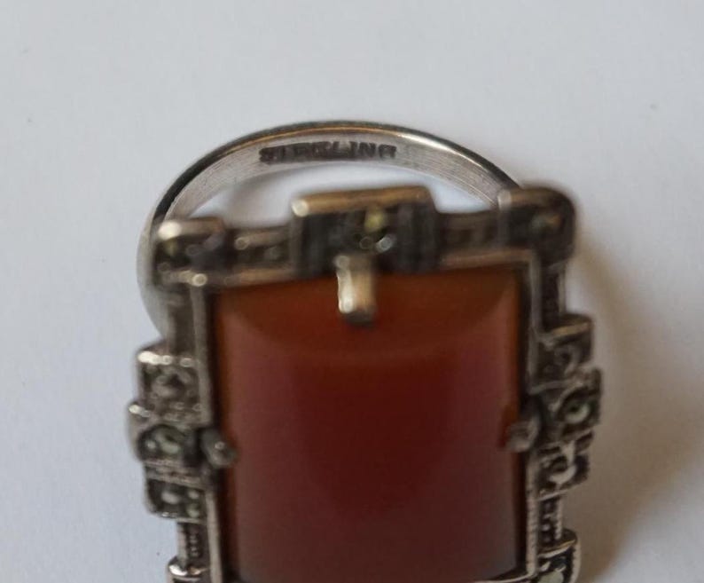 May include: A silver ring with a rectangular carnelian stone set in a marcasite frame. The ring is marked "STERLING".