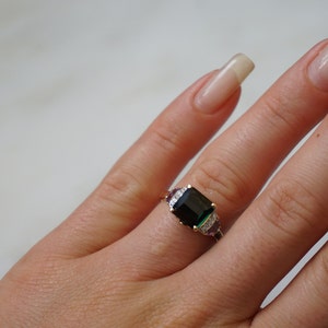 Pu&ograve; includere: Un anello d'oro con una grande pietra preziosa verde smeraldo circondata da pietre preziose pi&ugrave; piccole trasparenti e viola.
