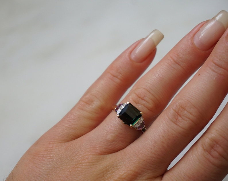 Pu&ograve; includere: Un anello d'oro con una grande pietra preziosa verde smeraldo circondata da pietre preziose pi&ugrave; piccole trasparenti e viola.