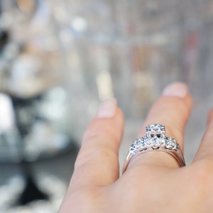 May include: A silver ring with a cluster of clear gemstones. The ring features a central square-cut stone flanked by smaller round stones. The band is a polished silver. The ring is on a person's finger.