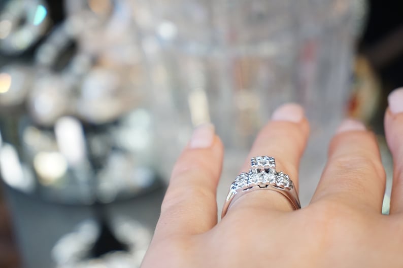 May include: A silver ring with a cluster of clear gemstones. The ring features a central square-cut stone flanked by smaller round stones. The band is a polished silver. The ring is on a person's finger.