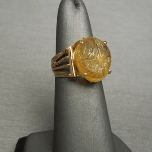 Op de afbeelding: Een gouden ring met een gegraveerde, ovale amber steen. De ringband heeft een touwachtig ontwerp.