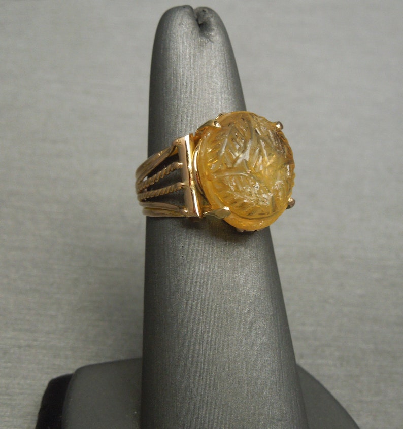 Op de afbeelding: Een gouden ring met een gegraveerde, ovale amber steen. De ringband heeft een touwachtig ontwerp.
