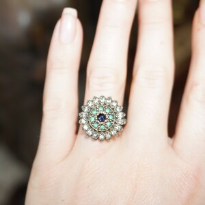 Pu&ograve; includere: Un anello d'argento con una pietra preziosa blu e verde circondata da pietre preziose pi&ugrave; piccole e trasparenti.
