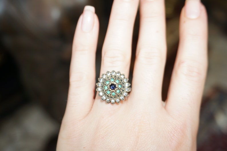 Pu&ograve; includere: Un anello d'argento con una pietra preziosa blu e verde circondata da pietre preziose pi&ugrave; piccole e trasparenti.