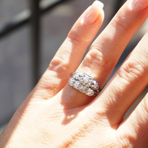 May include: A close-up of a diamond ring set on a finger. The ring features multiple small diamonds set in a cluster design. The band is a thin, silver-colored metal. The ring is worn on a hand with manicured nails.