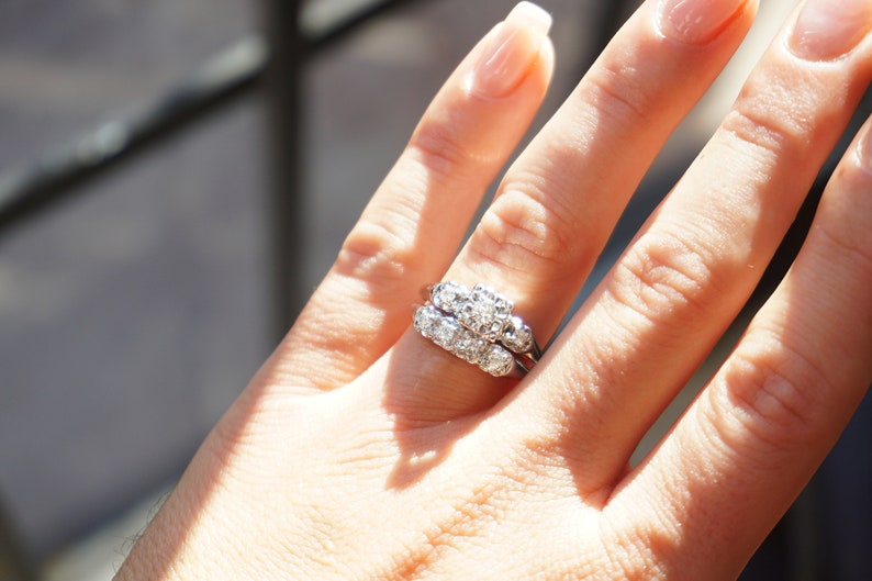 May include: A close-up of a diamond ring set on a finger. The ring features multiple small diamonds set in a cluster design. The band is a thin, silver-colored metal. The ring is worn on a hand with manicured nails.
