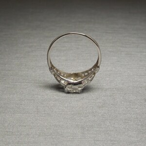 May include: A silver ring with a square-shaped center stone surrounded by smaller stones. The ring has a delicate filigree design.