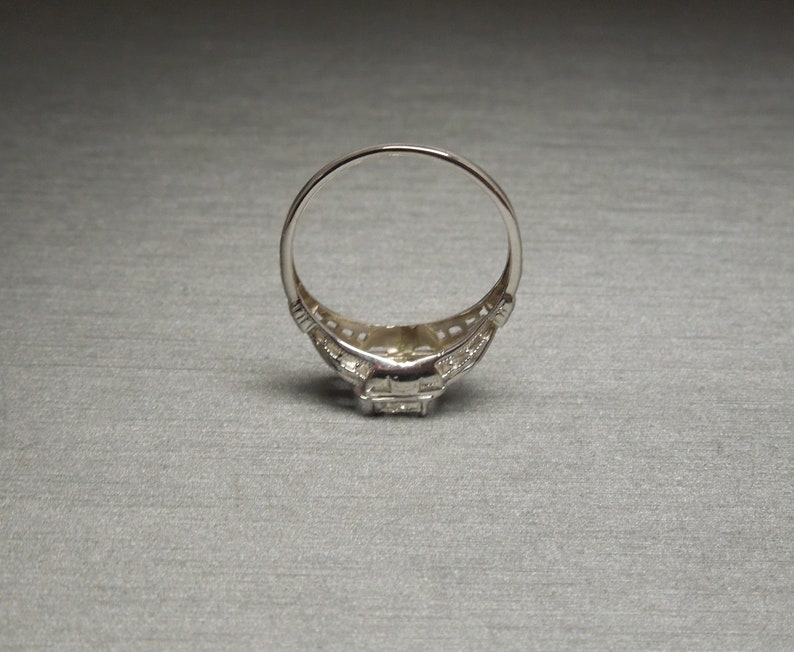 May include: A silver ring with a square-shaped center stone surrounded by smaller stones. The ring has a delicate filigree design.