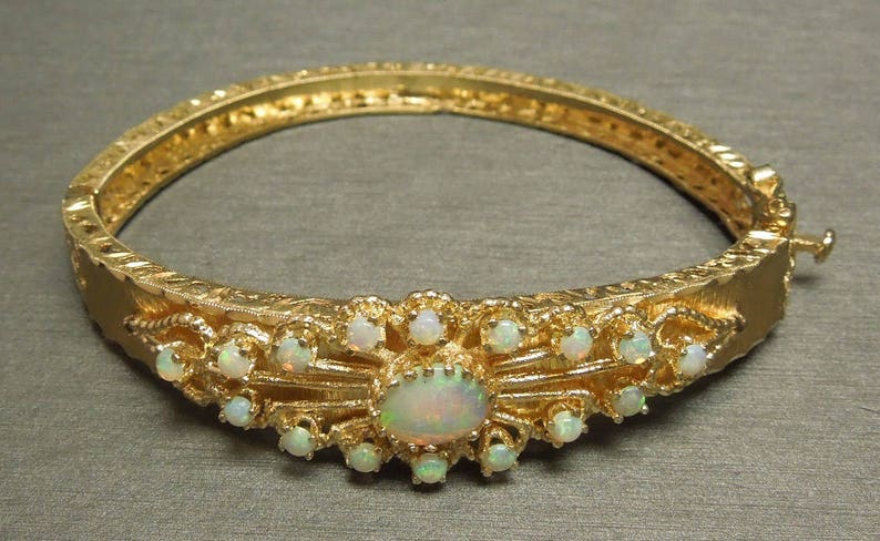 May include: A gold bangle bracelet with a delicate floral design and a large opal gemstone in the center. The bracelet is adorned with smaller opals around the center stone.