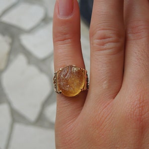 Op de afbeelding: Een gouden ring met een grote, ovale, gegraveerde, amberkleurige steen. De ring zit aan een vinger.