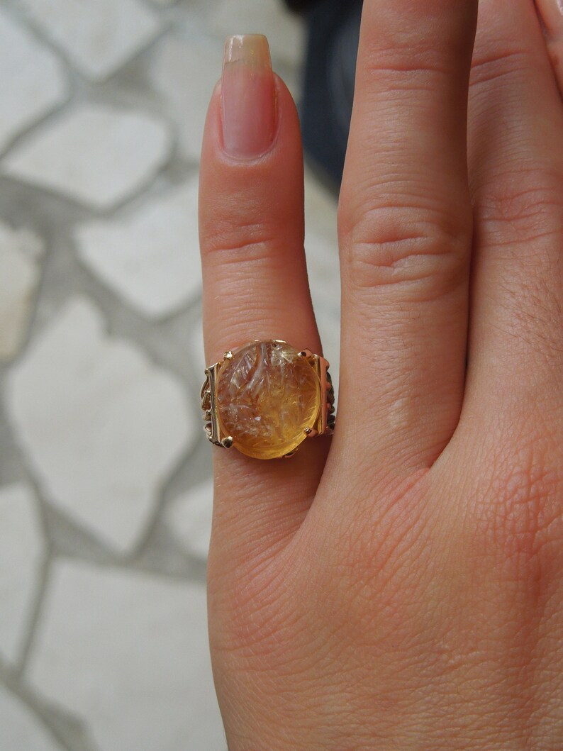 Op de afbeelding: Een gouden ring met een grote, ovale, gegraveerde, amberkleurige steen. De ring zit aan een vinger.