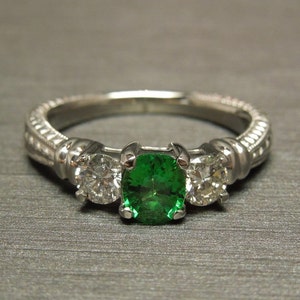 May include: A silver ring with a green gemstone and two smaller diamonds. The ring has a detailed band.