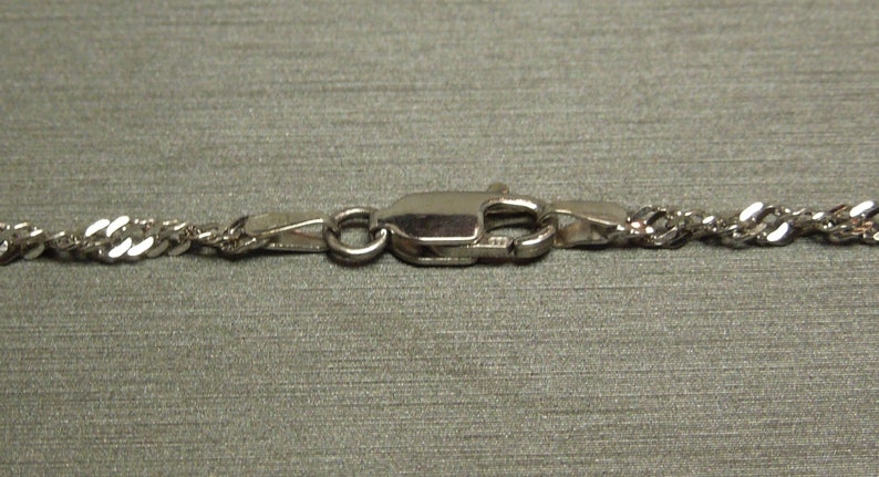K&ouml;nnte beinhalten: Nahaufnahme einer silberfarbenen Seilkette mit einem Karabinerverschluss. Die Kette hat ein gedrehtes Design, und der Verschluss ist sicher befestigt. Der Hintergrund ist ein neutrales Grau.