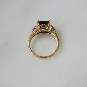 Pu&ograve; includere: Un anello d'oro con una grande pietra preziosa nera circondata da pietre preziose trasparenti pi&ugrave; piccole e due pietre preziose viola su entrambi i lati.