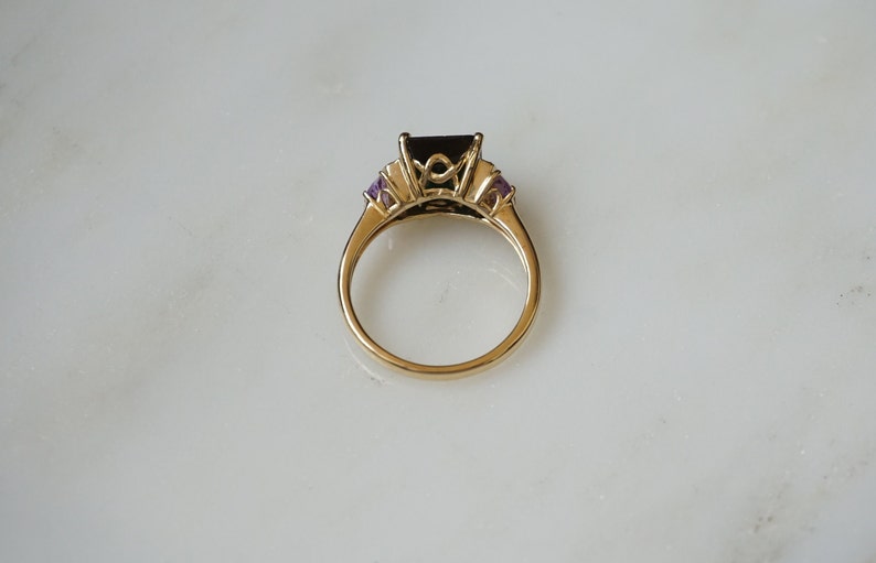 Pu&ograve; includere: Un anello d'oro con una grande pietra preziosa nera circondata da pietre preziose trasparenti pi&ugrave; piccole e due pietre preziose viola su entrambi i lati.