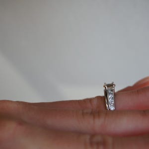 Pode incluir: Um anel de noivado de prata com um grande diamante de forma retangular e diamantes menores engastados em uma banda.