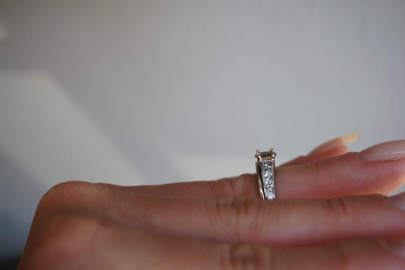 Pode incluir: Um anel de noivado de prata com um grande diamante de forma retangular e diamantes menores engastados em uma banda.