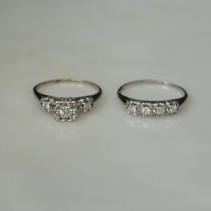 May include: Two silver rings with small, sparkling gemstones. One ring features three stones clustered together, while the other has a row of five stones. The rings are set against a white background.