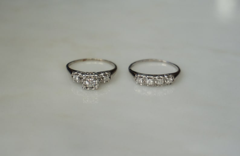 May include: Two silver rings with small, sparkling gemstones. One ring features three stones clustered together, while the other has a row of five stones. The rings are set against a white background.