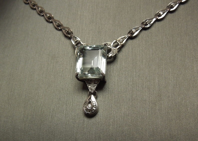 K&ouml;nnte beinhalten: Elegante Silberkette mit einem gro&szlig;en, rechteckigen, hellblauen Edelsteinanh&auml;nger. Der Anh&auml;nger ist mit kleineren Diamanten und einem tropfenf&ouml;rmigen Anh&auml;nger verziert. Die Kette hat ein filigranes Design mit kleinen Diamantakzenten.