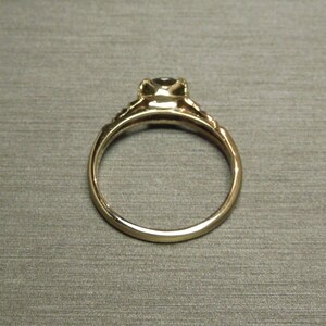 May include: A gold ring with a single, round, dark gemstone set in a prong setting. The ring has a delicate, ornate band.