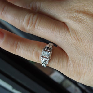May include: A silver ring with a clear, rectangular-shaped gemstone set in a delicate, intricate design.