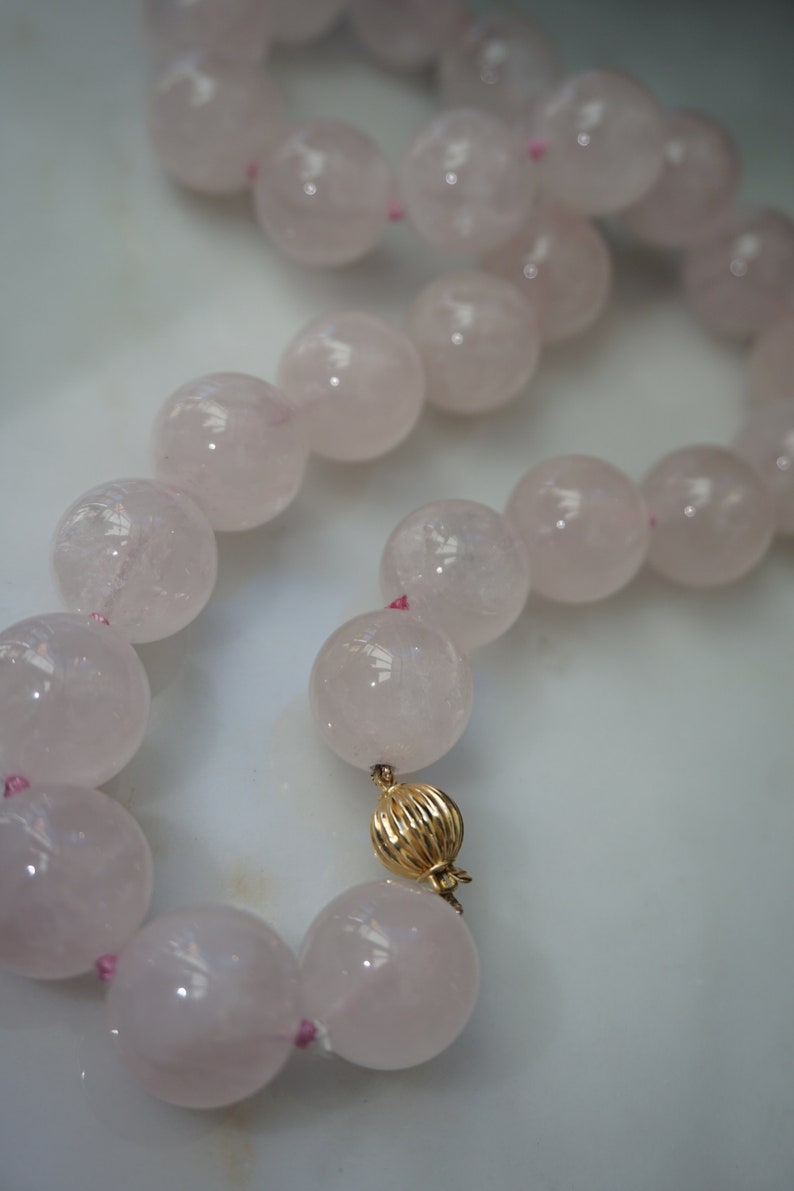 May include: A pink rose quartz beaded necklace with a gold clasp. The beads are large and round, and the necklace is strung on a pink thread.
