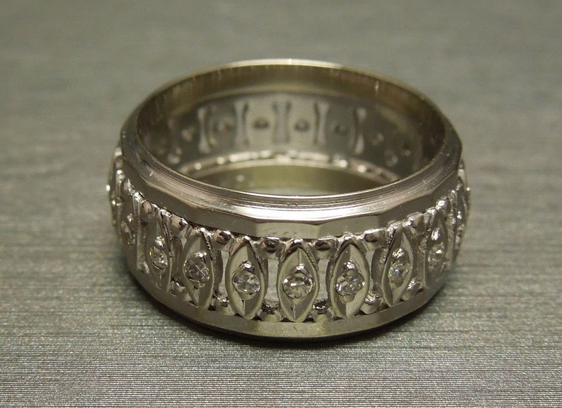 May include: A silver wedding band with a decorative design featuring small diamonds set in a repeating pattern.