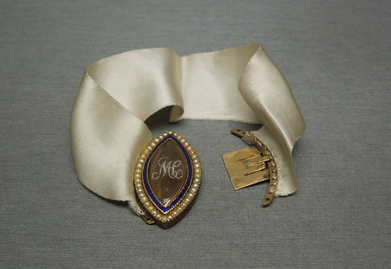 May include: A gold locket with a blue enamel border and pearl accents. The locket is open and shows a hair lock with the initials "MC" inscribed. The locket is attached to a white satin ribbon with a gold clasp.