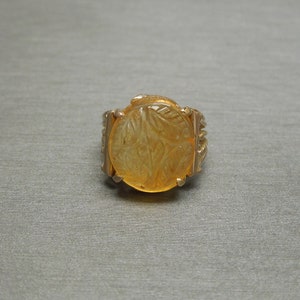 Op de afbeelding: Een gouden ring met een grote, ronde, gele edelsteen. De edelsteen heeft een gegraveerd ontwerp.