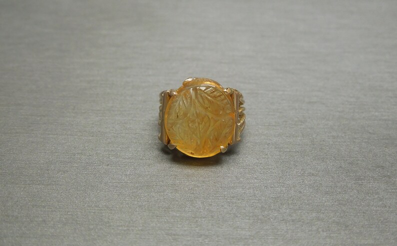 Op de afbeelding: Een gouden ring met een grote, ronde, gele edelsteen. De edelsteen heeft een gegraveerd ontwerp.
