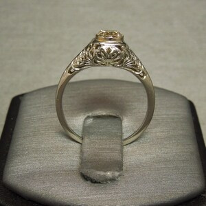 Puede incluir: Un delicado anillo de compromiso de filigrana con un solo diamante redondo engastado en una banda de oro blanco.