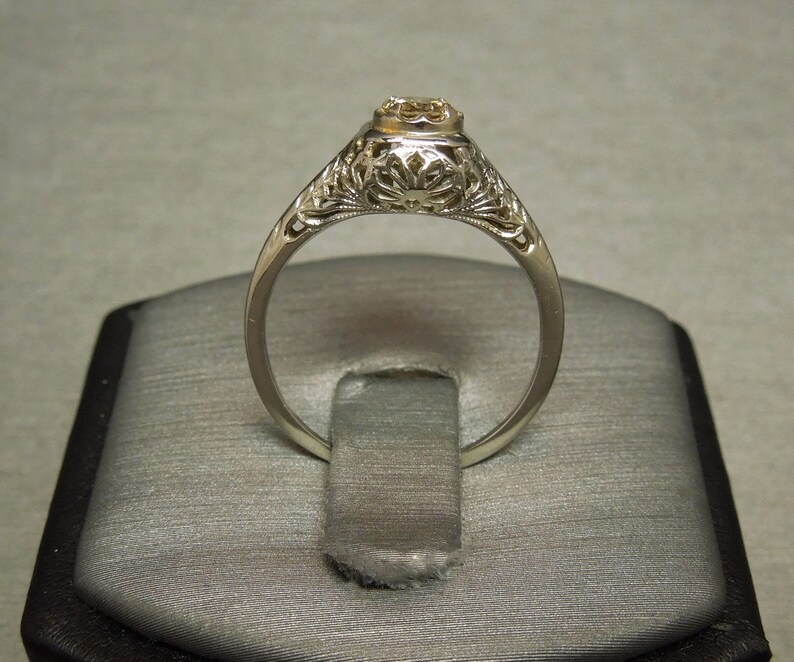 Puede incluir: Un delicado anillo de compromiso de filigrana con un solo diamante redondo engastado en una banda de oro blanco.