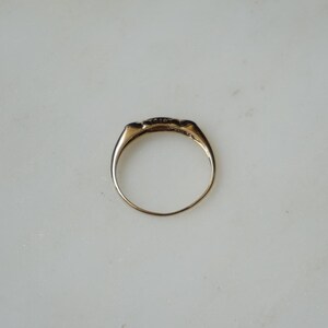 May include: A gold ring with a rectangular face, set with small, dark stones. The ring has a smooth, polished band and a classic design. The ring is shown on a white surface.