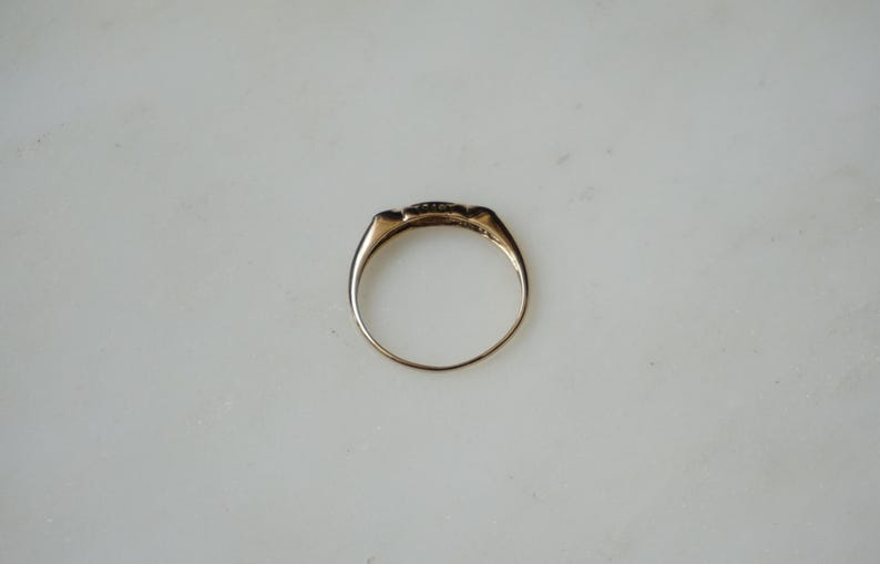 May include: A gold ring with a rectangular face, set with small, dark stones. The ring has a smooth, polished band and a classic design. The ring is shown on a white surface.