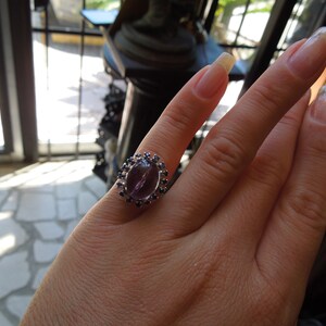 May include: A silver ring featuring a large, oval-shaped, purple gemstone as the centerpiece. The gemstone is surrounded by a halo of smaller, dark blue gemstones. The ring is displayed on a hand.