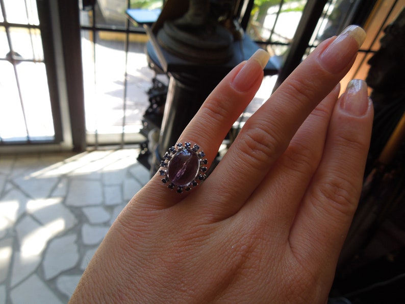 May include: A silver ring featuring a large, oval-shaped, purple gemstone as the centerpiece. The gemstone is surrounded by a halo of smaller, dark blue gemstones. The ring is displayed on a hand.