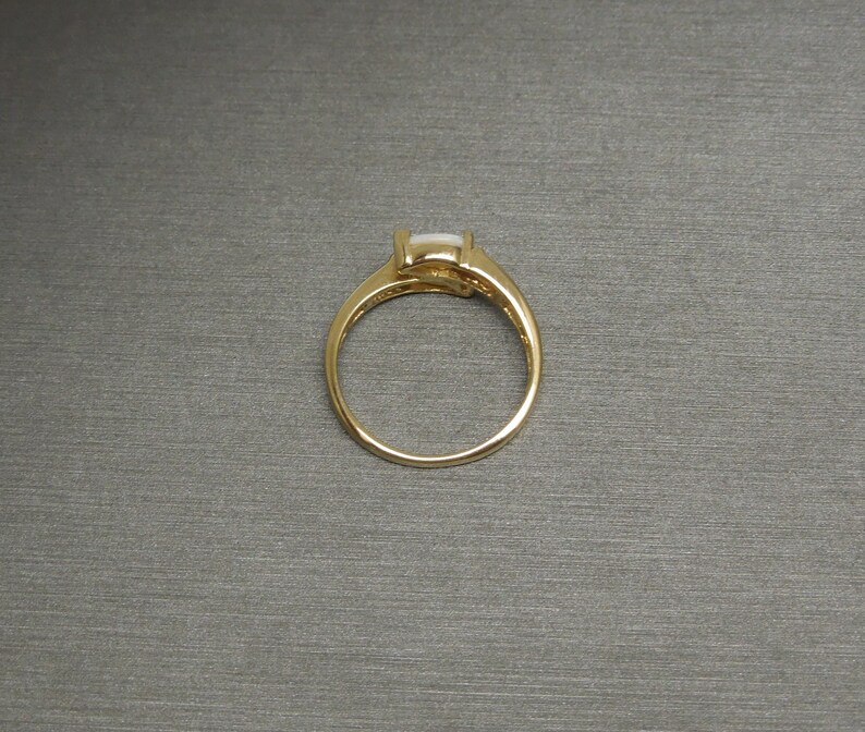 K&ouml;nnte beinhalten: Ein goldener Ring mit einem wei&szlig;en ovalen Edelstein, der in einer Krappenfassung sitzt.