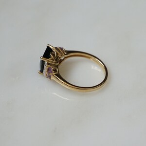 Pu&ograve; includere: Un anello d'oro con una grande pietra preziosa nera circondata da pietre preziose pi&ugrave; piccole trasparenti e viola.