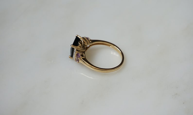 Pu&ograve; includere: Un anello d'oro con una grande pietra preziosa nera circondata da pietre preziose pi&ugrave; piccole trasparenti e viola.